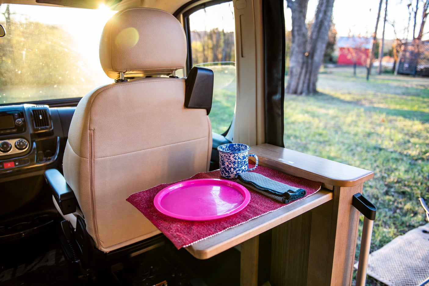 Cozy RV rental interior: passenger seat, pop-out table with pink plate, mug. Sunny view of campsite.