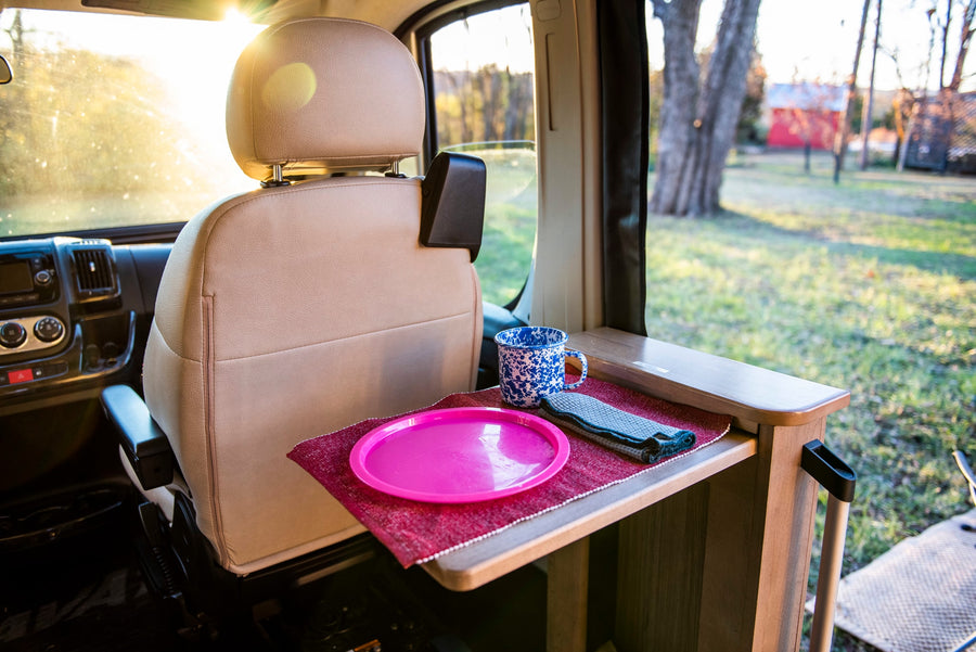 Cozy RV rental interior: passenger seat, pop-out table with pink plate, mug. Sunny view of campsite.