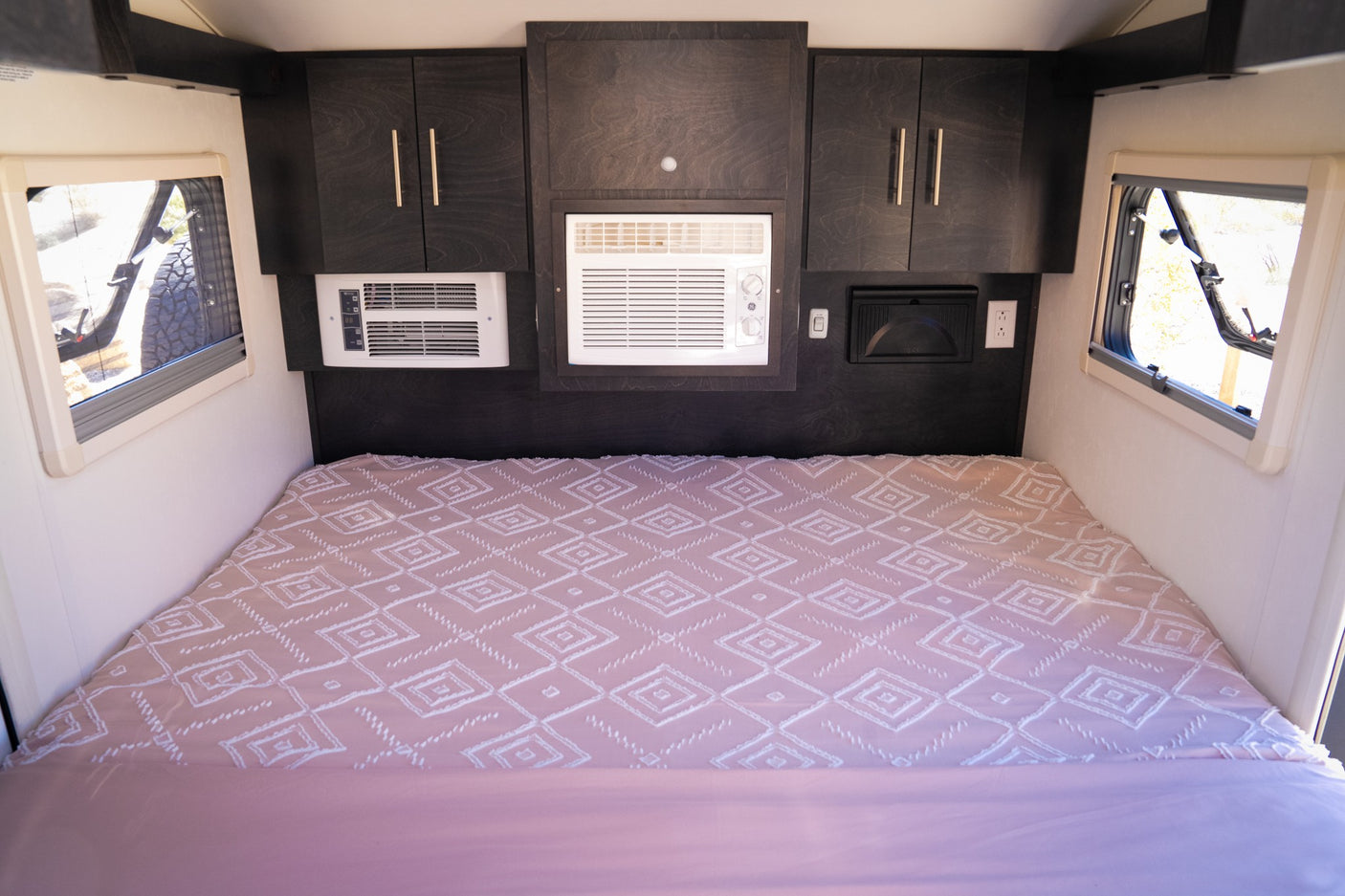 Inside an RV rental: a bed with pink patterned bedding, flanked by windows, under dark wood cabinets & AC units.