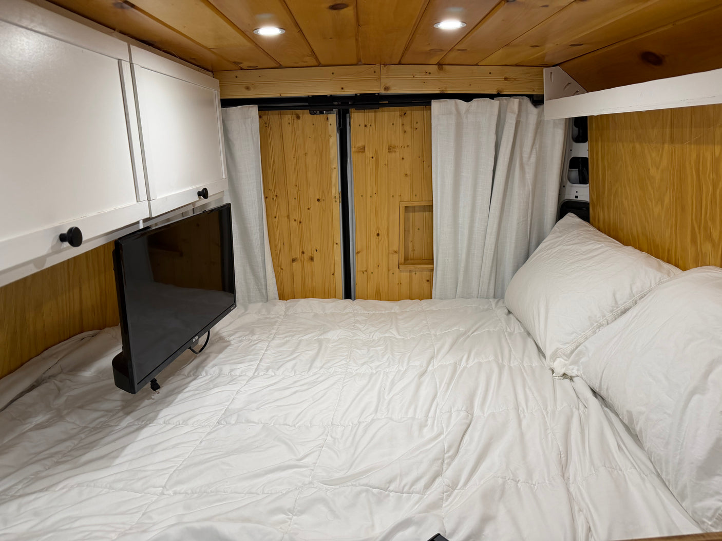 Comfortable RV rental van interior features a white bed, TV, overhead cabinets, and warm wood finishes.