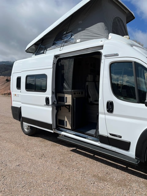 A white camper van RV rental with its pop-top roof extended and side door open, revealing a kitchenette and seating.