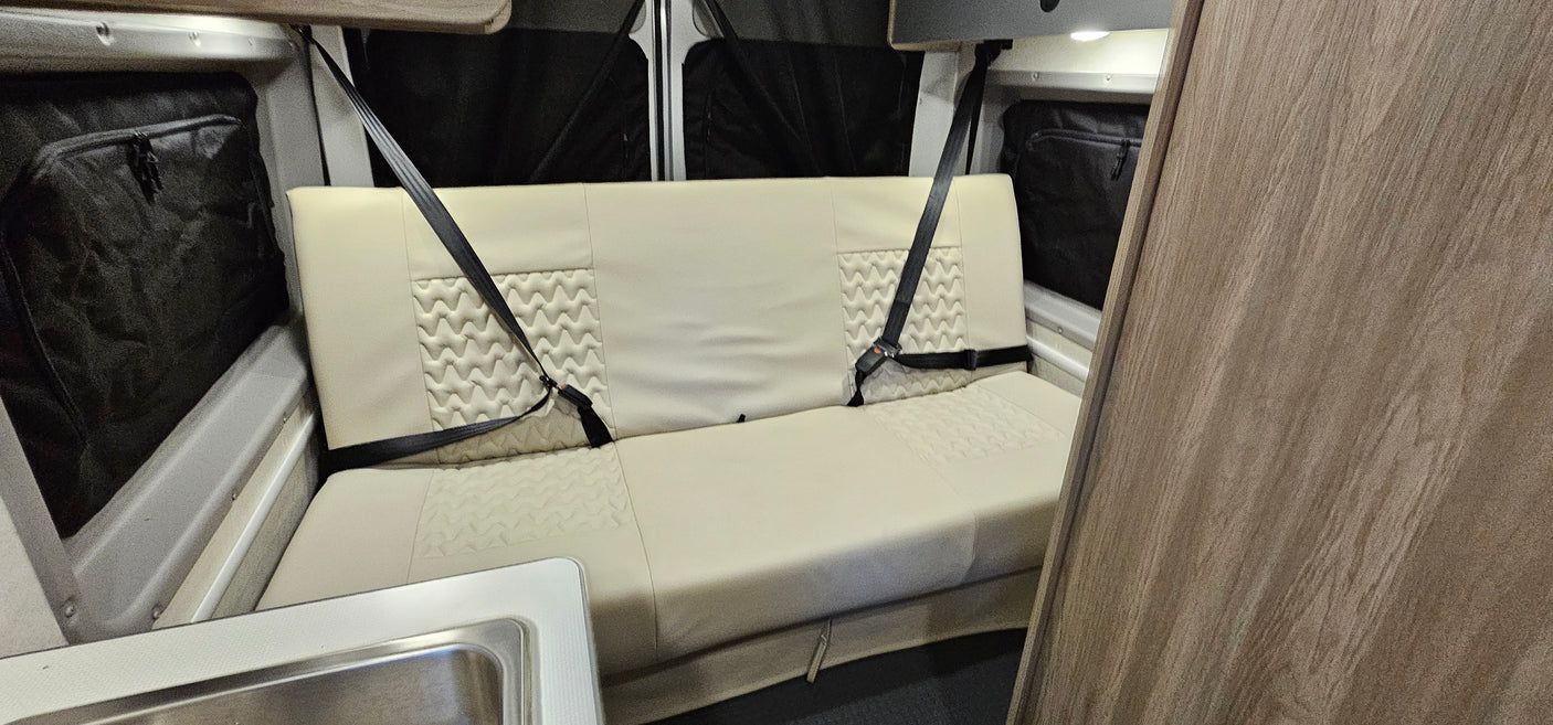 An rv rental's cream bench seat with two seatbelts. Features quilted design, sink, and window storage.