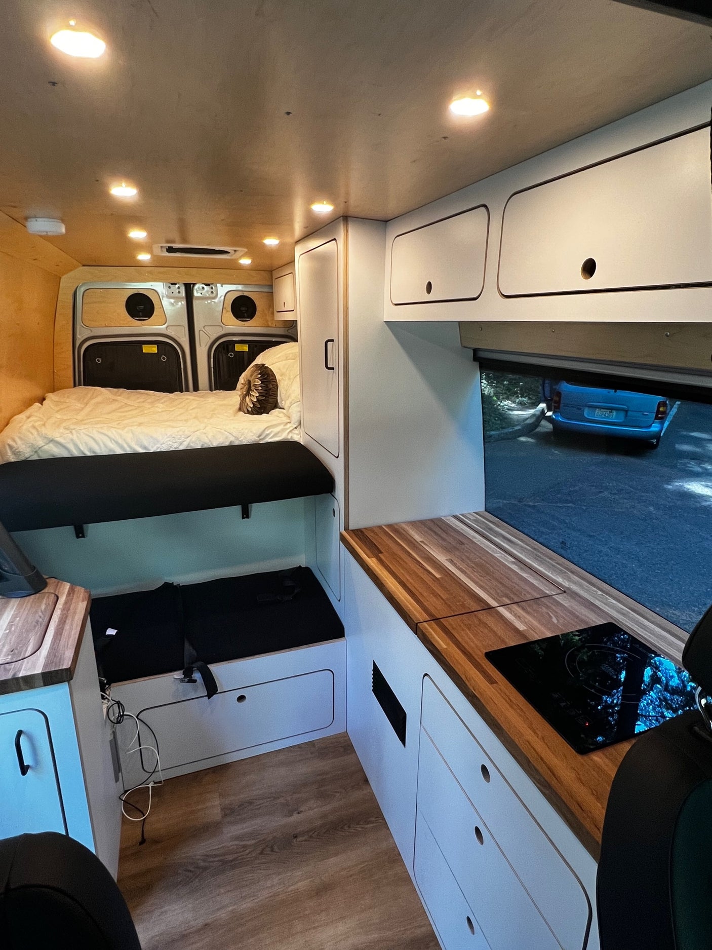 Modern RV rental interior with cozy bed, kitchen cooktop, custom storage, and view of a car on the road outside.