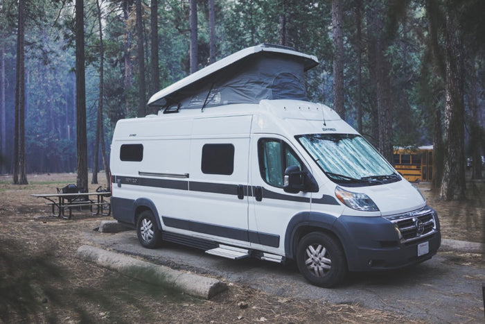A white pop-top RV rental camper van parked on pavement in a dense forest campsite. A picnic table is nearby.