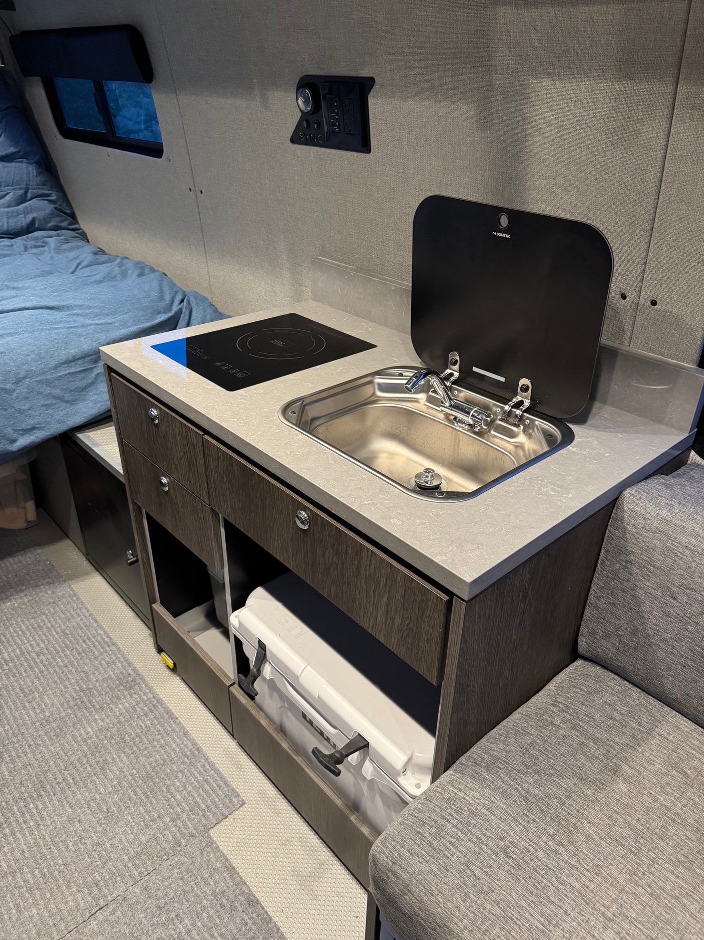 RV rental: Compact kitchen with induction cooktop, sink, and cooler storage. Includes a bed and seating for mobile living comfort.