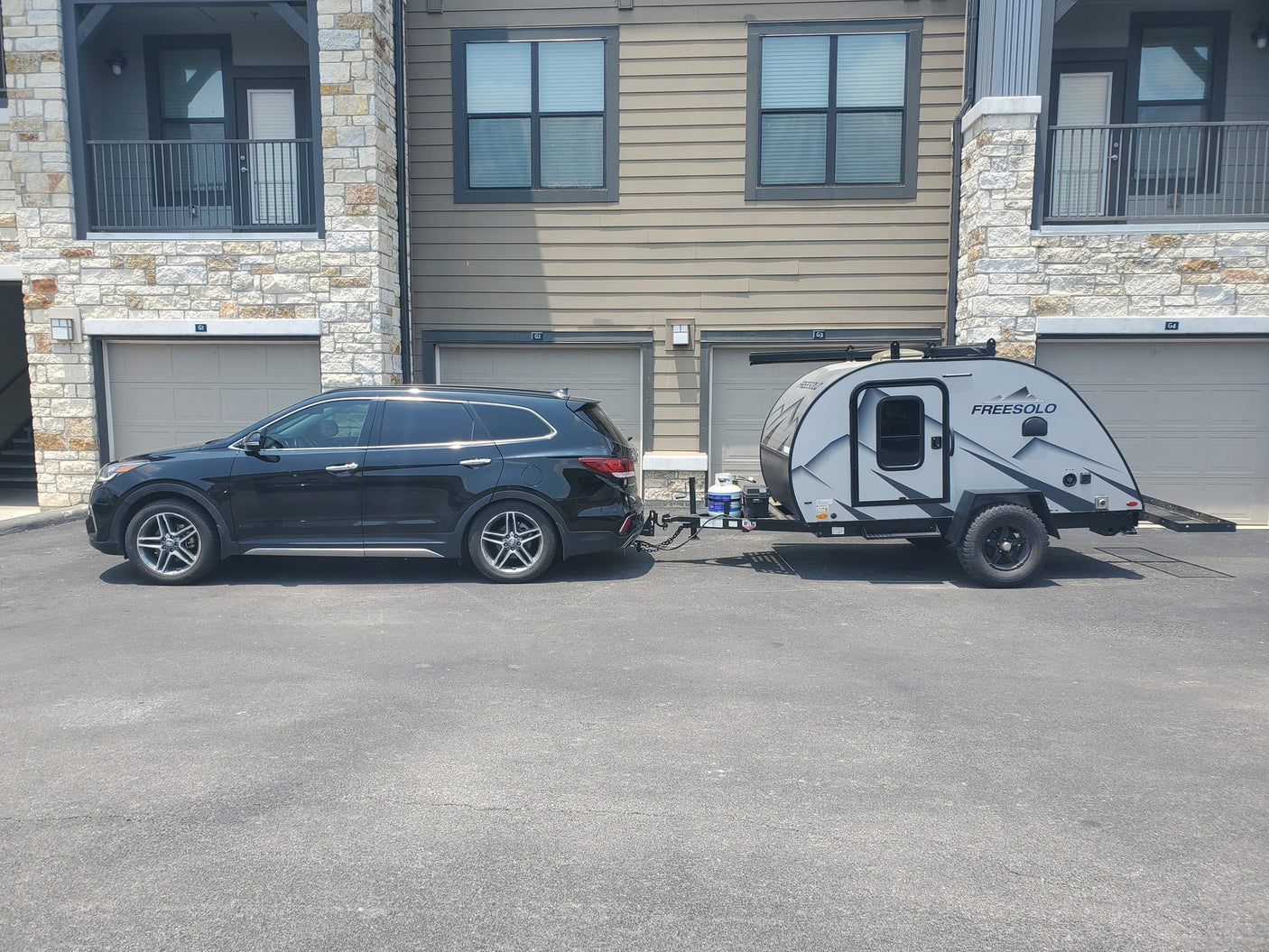 A black SUV is hitched to a grey teardrop RV rental, a FreeSolo camper, parked outside apartments.