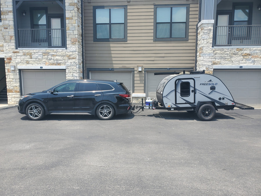 A black SUV is hitched to a grey teardrop RV rental, a FreeSolo camper, parked outside apartments.