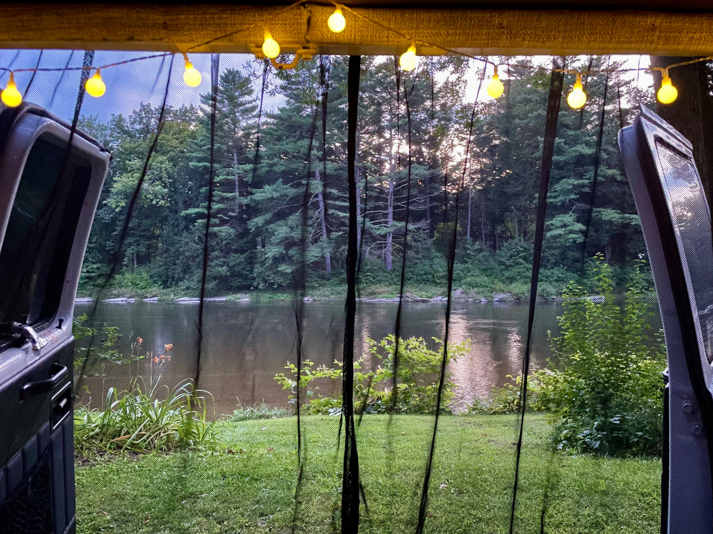 Serene river and forest view from an RV rental, seen through a mesh screen with string lights overhead. Perfect outdoor escape.