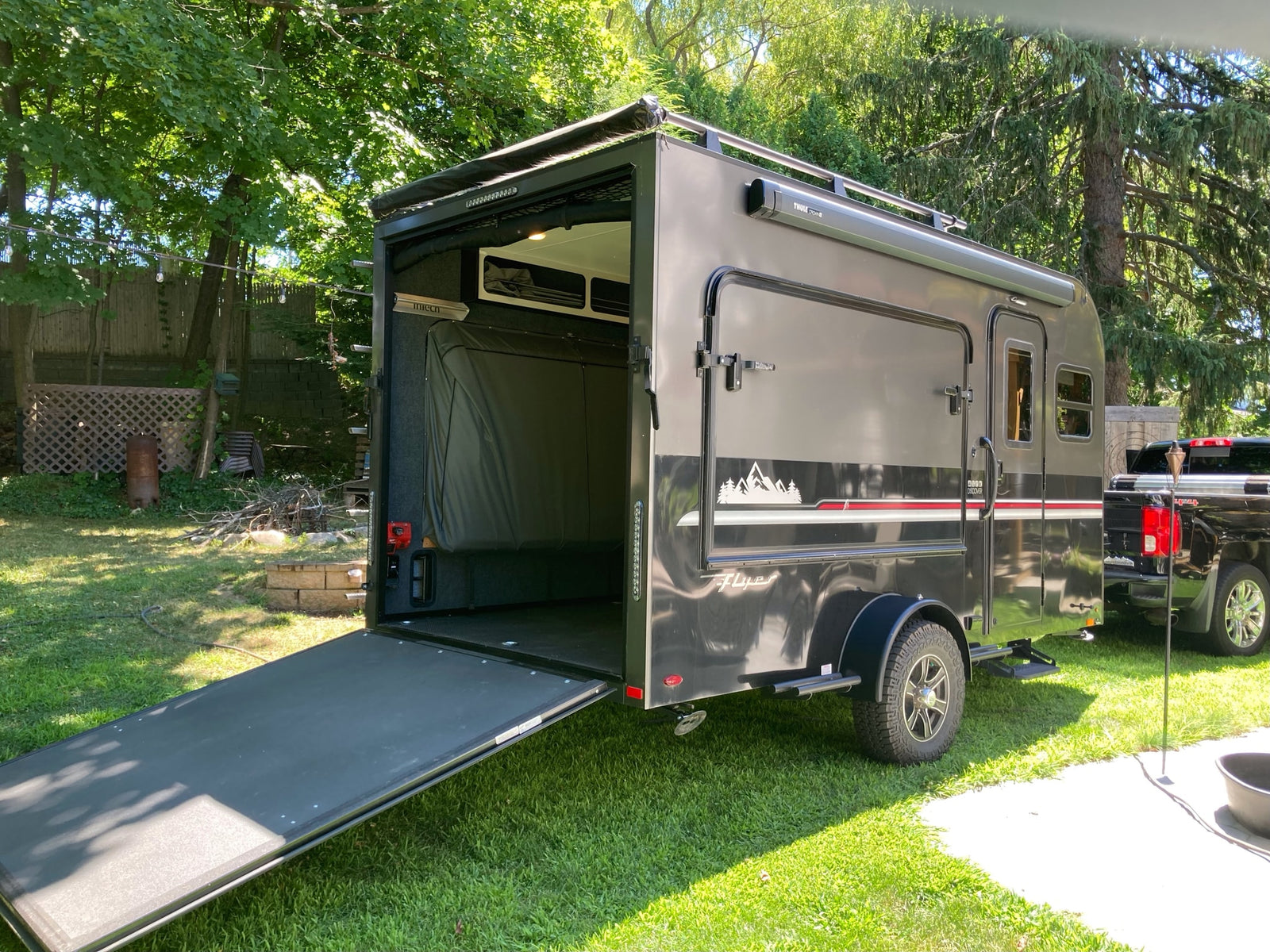 This grey adventure rv rental trailer has an open ramp door, fold-up bed inside, Thule awning, & rugged tires.