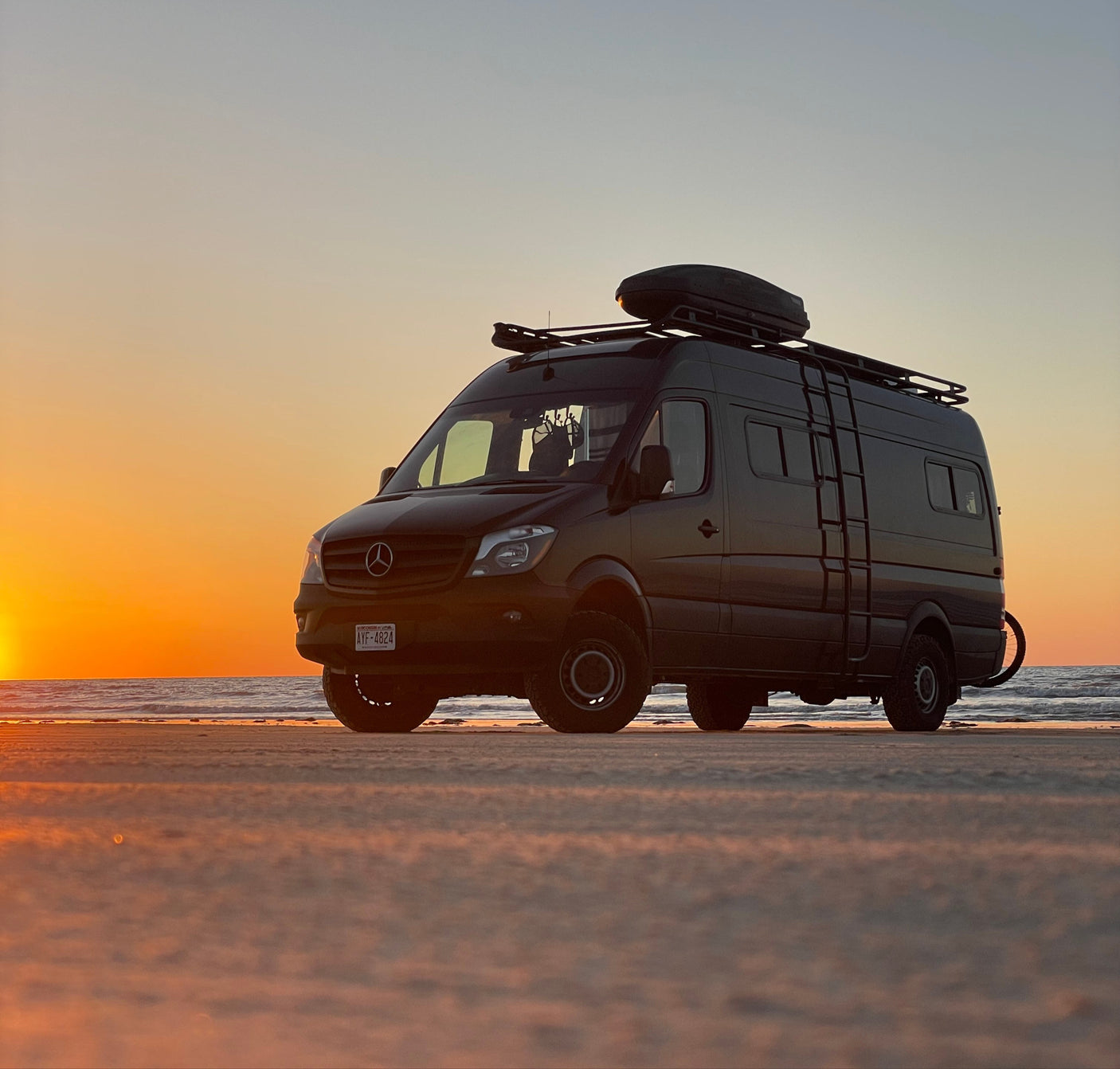 A rugged Mercedes Sprinter RV rental van silhouetted against a vibrant sunset on a sandy beach, ready for adventure.
