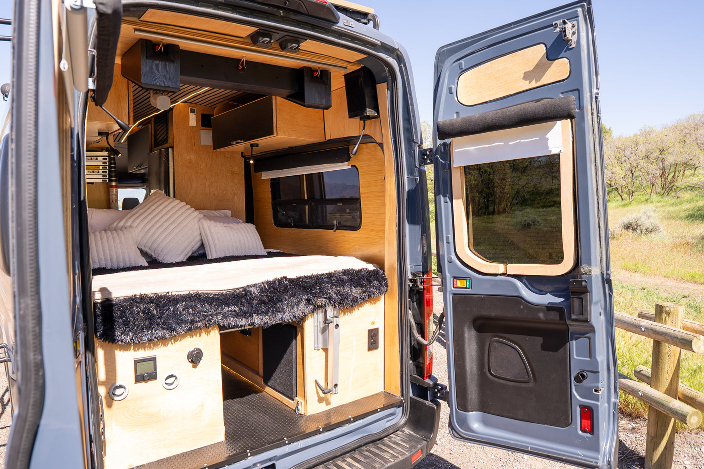 Open rear doors of an RV rental camper van reveal a custom wooden interior with a bed, storage, and a scenic outdoor view.
