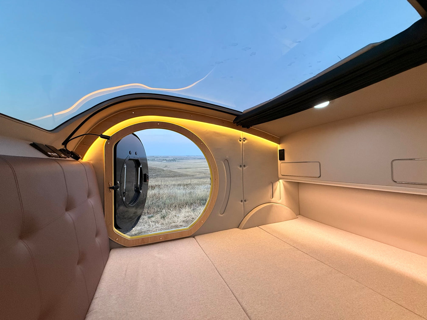 Modern RV rental interior: spacious bed, large panoramic skylight, and circular window revealing a vast, open landscape.