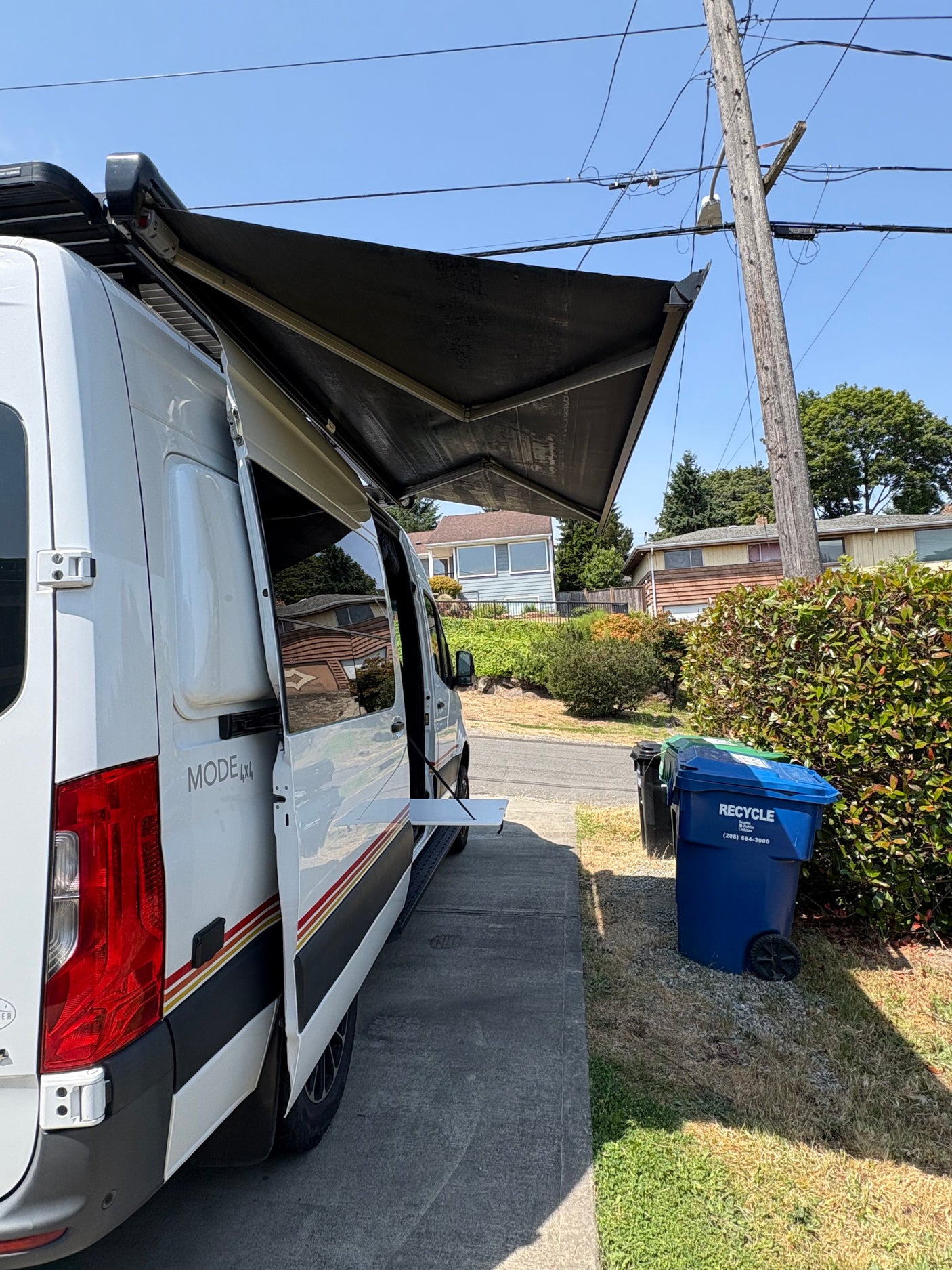 A white RV rental van with an extended awning and open side door sits on a sunny driveway beside a blue recycling bin.