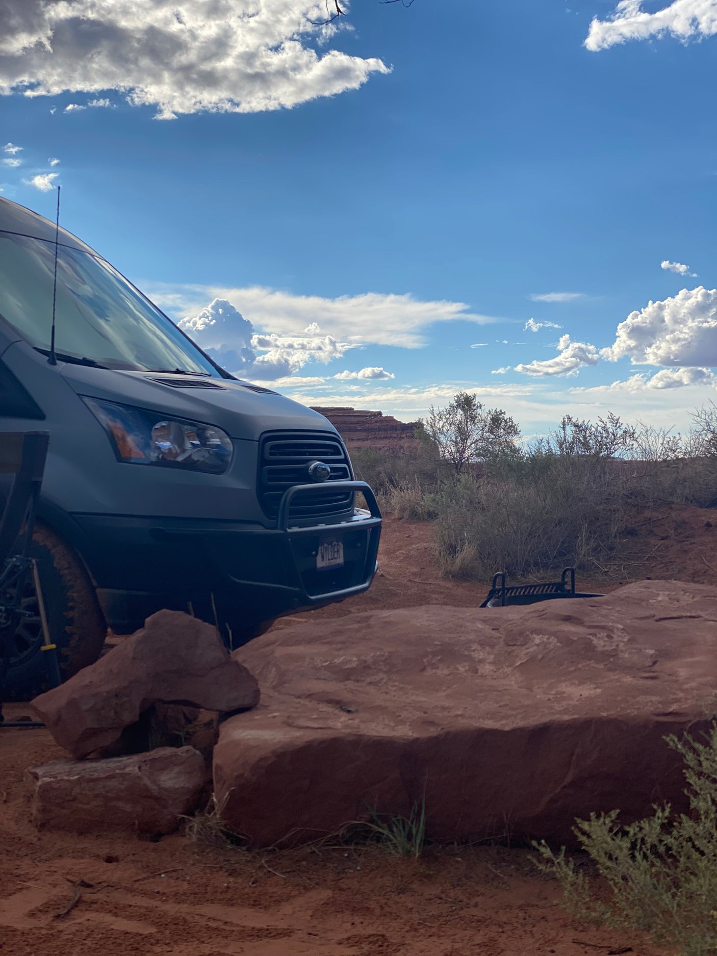 A gray Ford Transit van, an rv rental, is parked on a dirt path amidst red rock formations and desert shrubs under a cloudy sky.