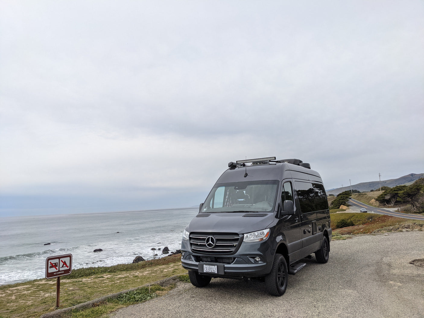 Dark gray Mercedes Sprinter RV rental parked at an ocean overlook on a cloudy day, with a scenic coastal road curving uphill.