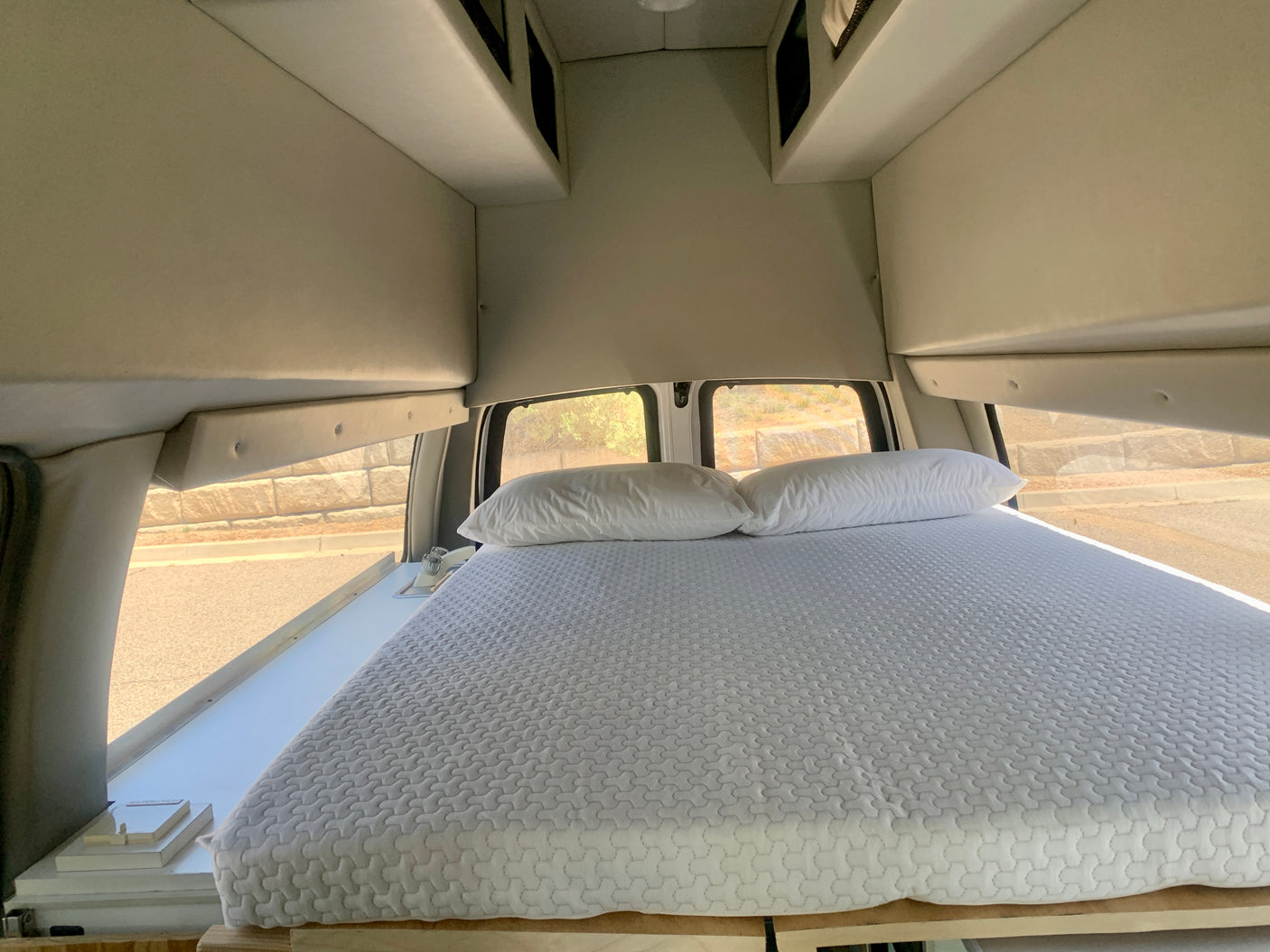 Inside this cozy RV rental camper van: a bed with white pillows, overhead storage, and windows revealing the road outside.