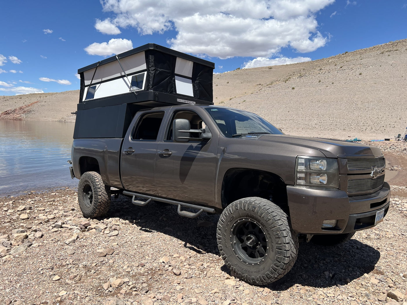 Oru Designs Bruin pop-up truck camper with the roof raised and side windows open, mounted on a lifted Chevrolet Silverado 2500HD parked on a rocky desert lakeshore — photo 1