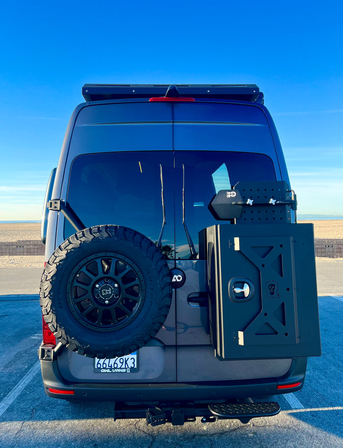 Rear of a gray RV rental van with a spare tire and storage box. Parked at a sunny beach, ready for adventure.