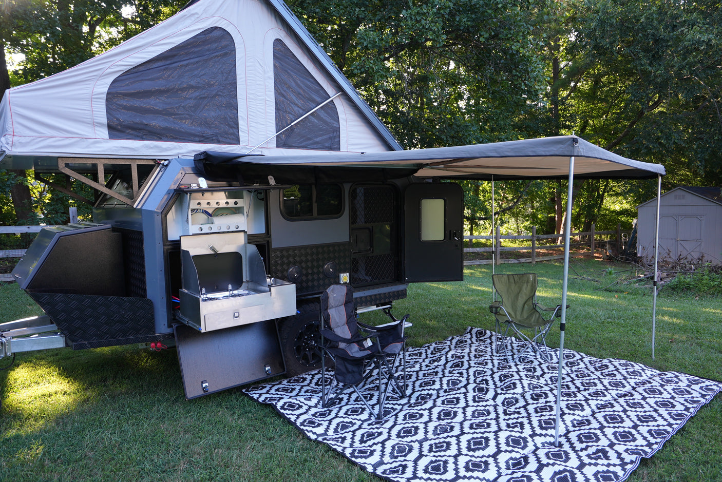 This grey and black rv rental pop-up camper is set up outdoors with an extended awning, outdoor kitchen, chairs, and mat.