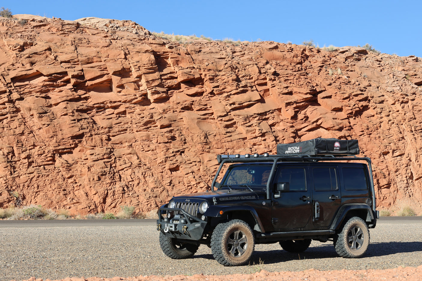 Black Jeep Wrangler with roof tent, an adventure RV rental, parked roadside against towering red rock formations under blue sky.
