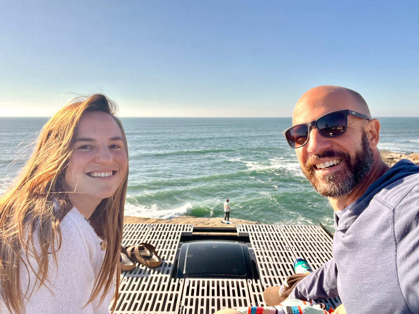 Smiling couple on their RV rental rooftop, enjoying vast ocean views and the coastal breeze, with a person by the waves below.