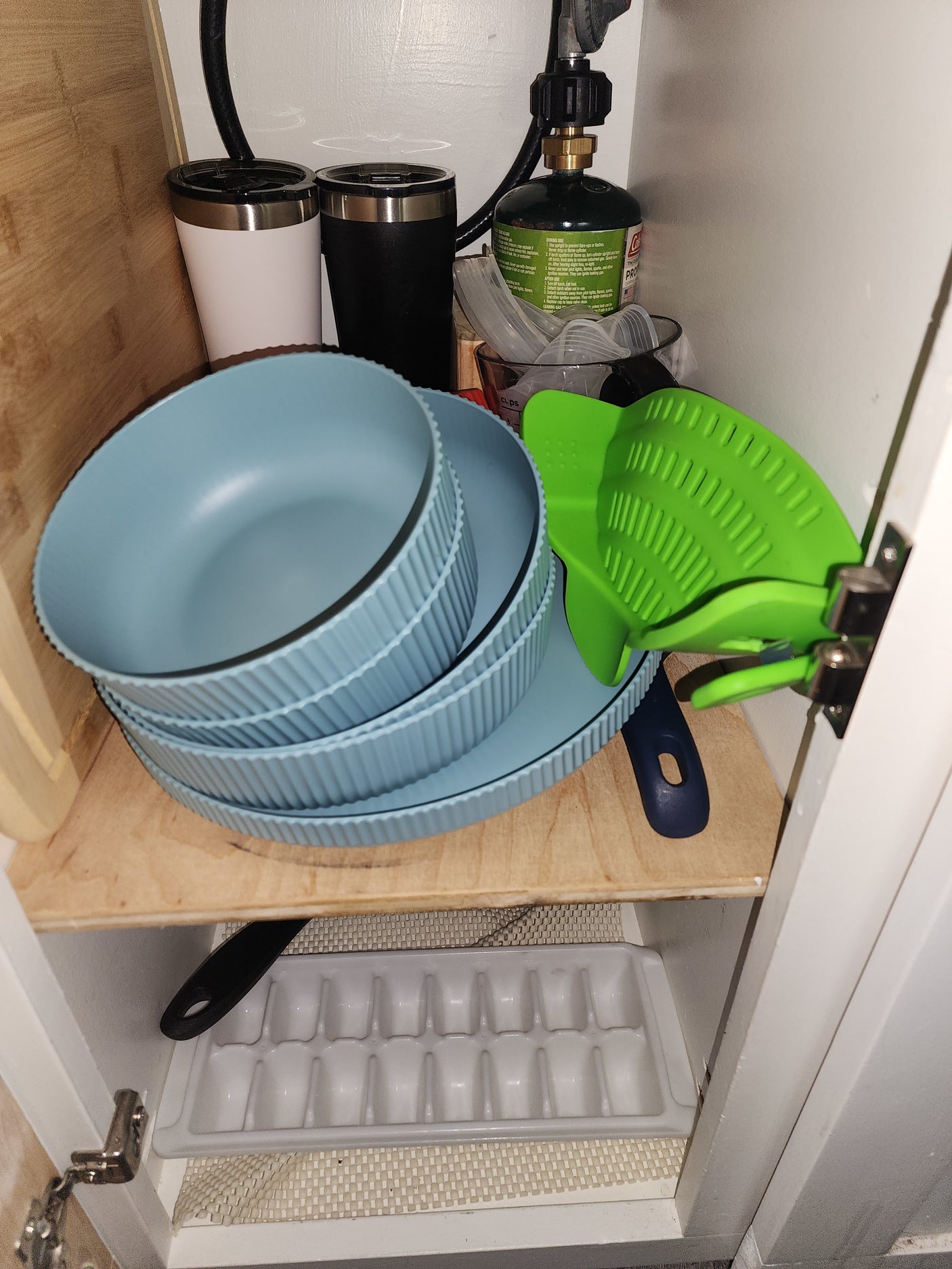 Inside an RV rental cabinet: stacked blue dishes, a green propane tank, travel mugs, and a bright green silicone strainer.