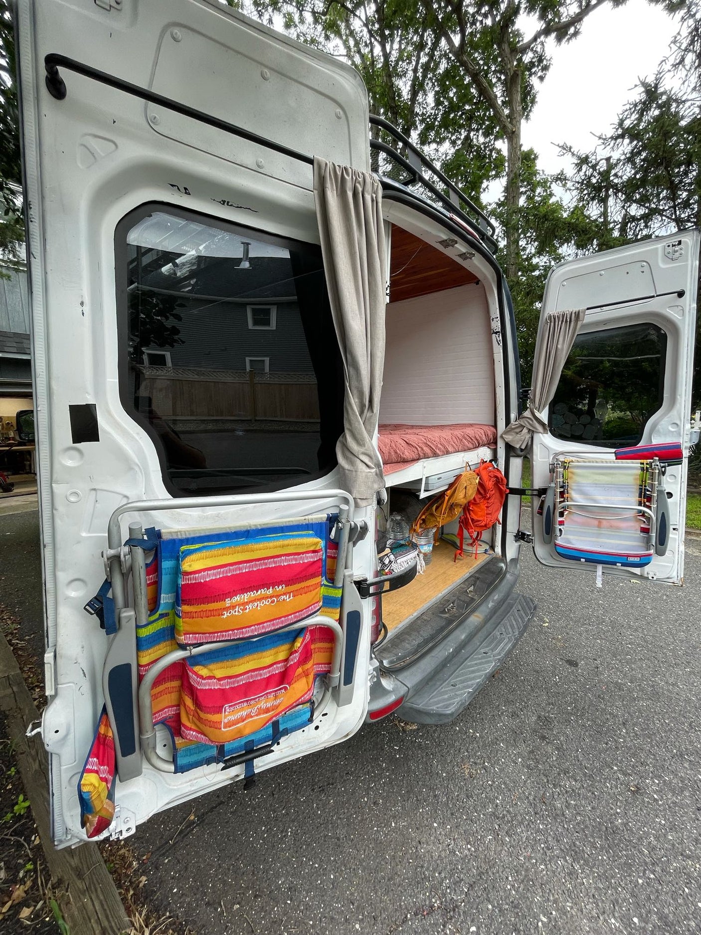 Open white camper van rear, revealing a bed, stored gear, and door-mounted beach chairs/drying rack. Great for an RV rental trip.