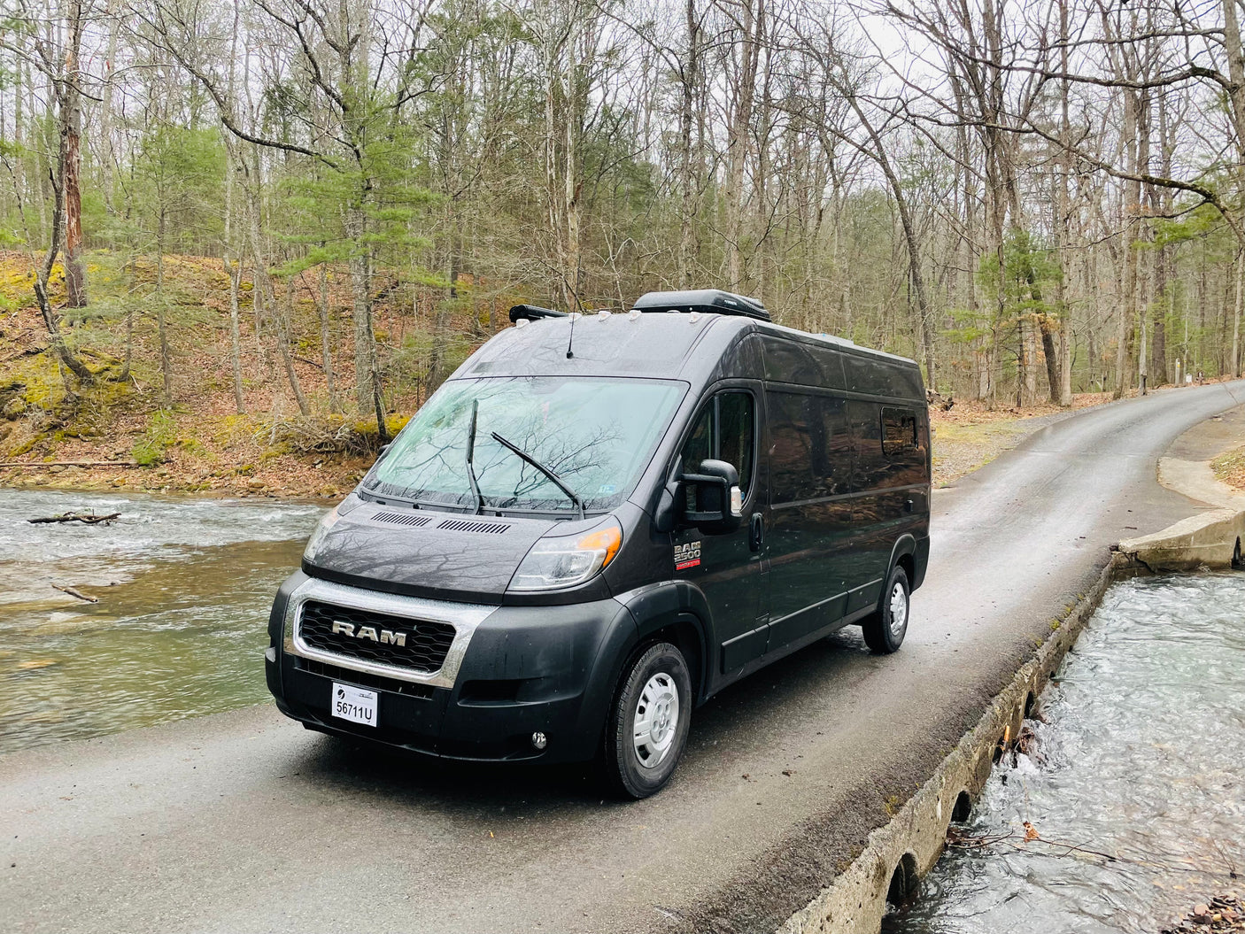 An RV rental, a grey Ram ProMaster camper van, is parked on a bridge over a stream in a serene forest setting.