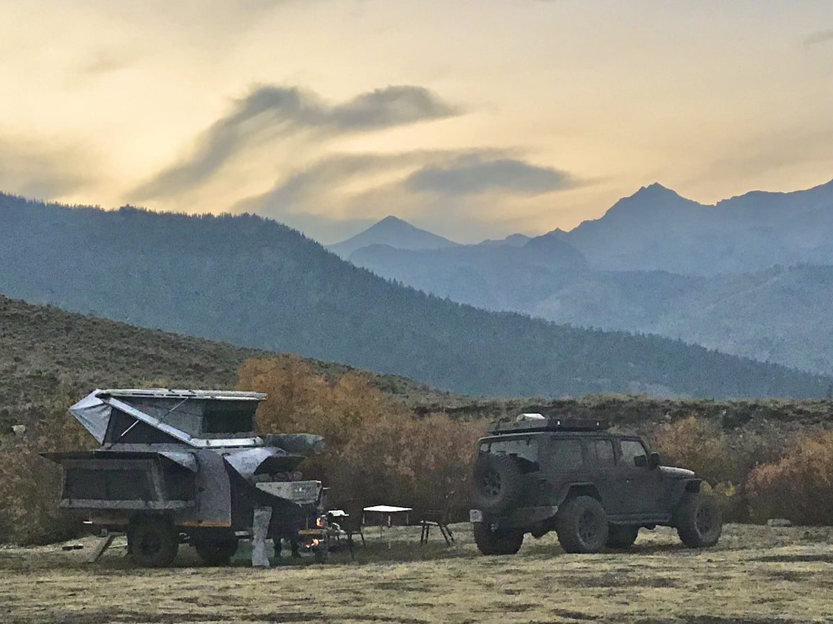 Dusk camp in an open mountain valley with the rooftop tent raised, awning out and a campfire burning beside the trailer, layered ridgelines behind — photo 5