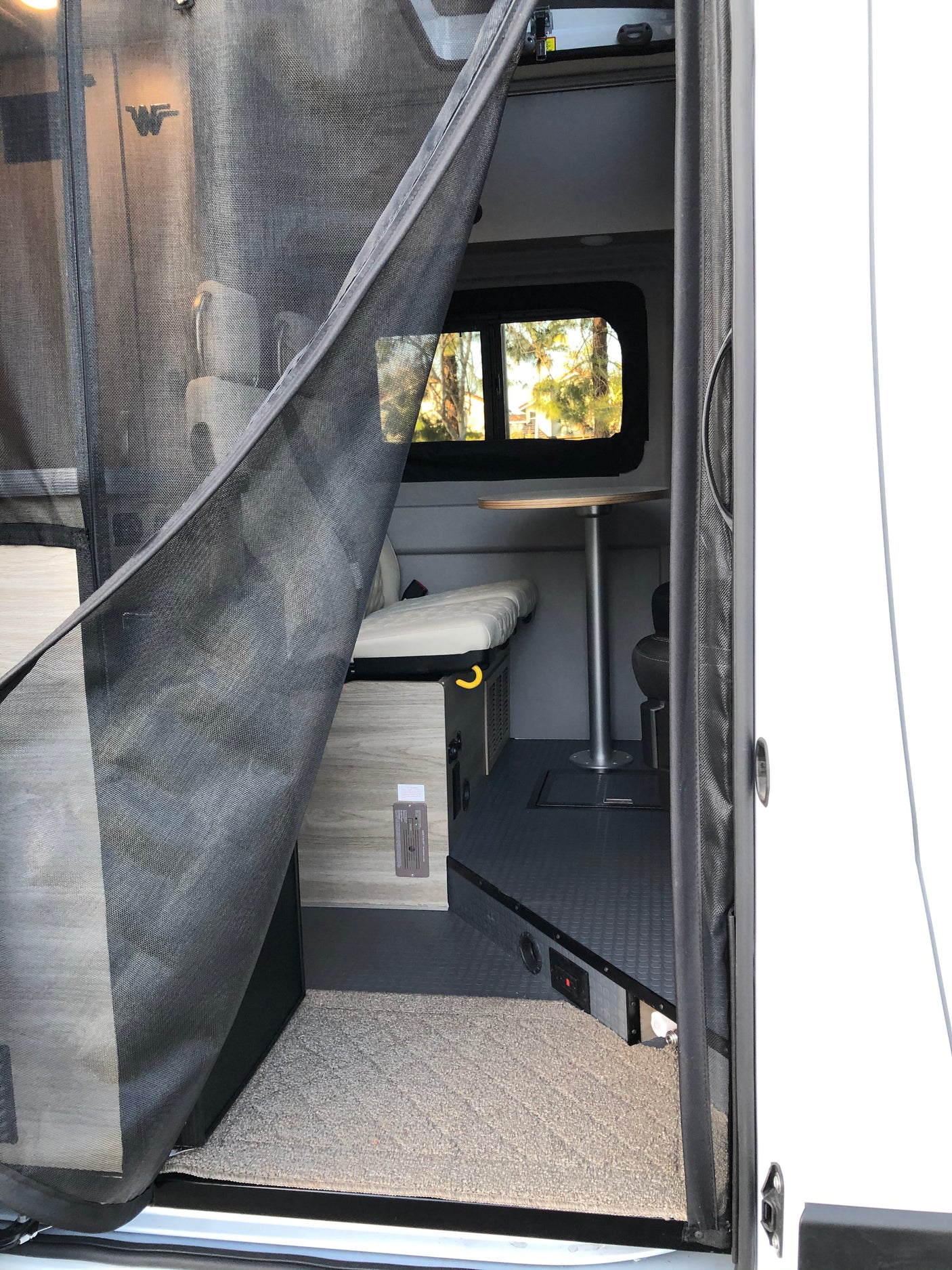 Inside a modern RV rental: open screen door reveals a functional living space with a dinette, bench, and window view.