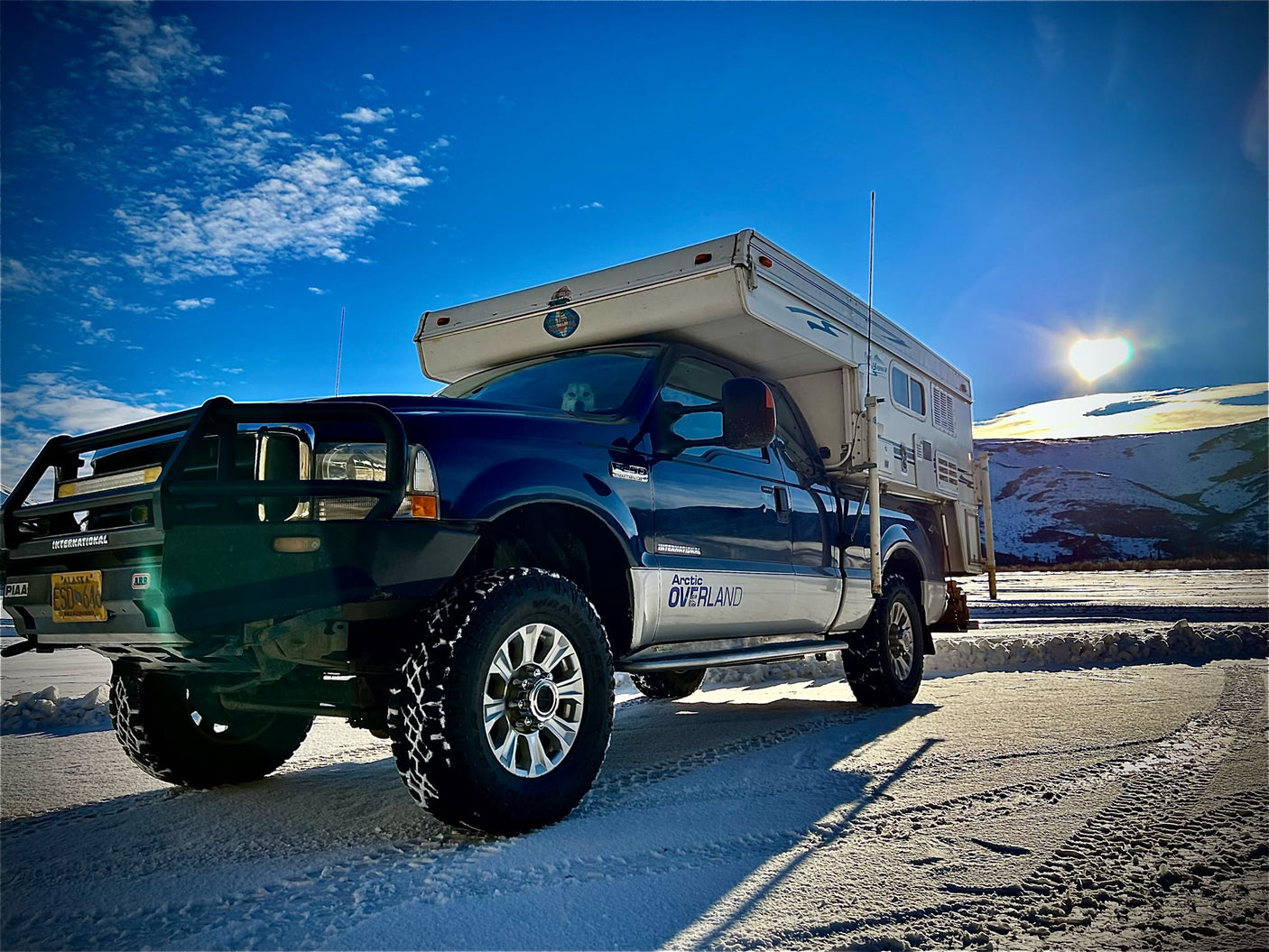 Blue Ford truck with Arctic Overland camper on snow. Heart-shaped sun. Perfect RV rental for Alaska winter trips with your dog.|Blue truck with Bronco pop-up truck camper