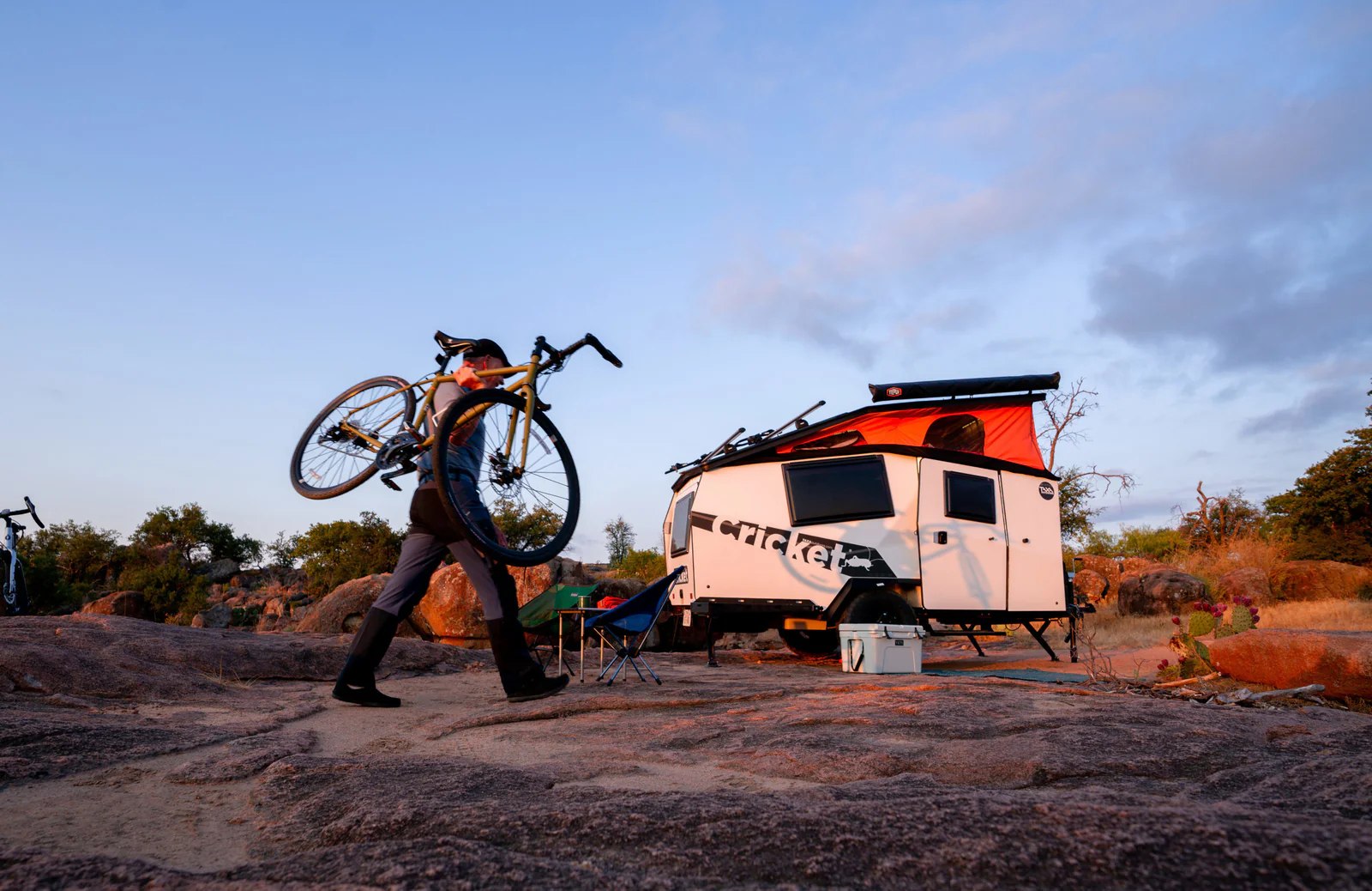 Person carrying a bike on rocky terrain next to a white RV rental camper, set up for outdoor adventure.