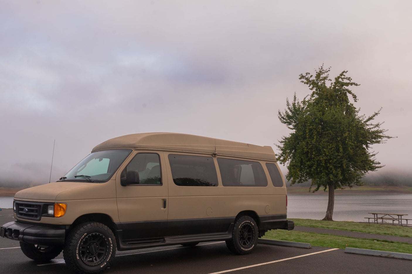 Tan Ford E-Series camper van (RV rental) parked in a lot beside a foggy lake with a tree & picnic area.