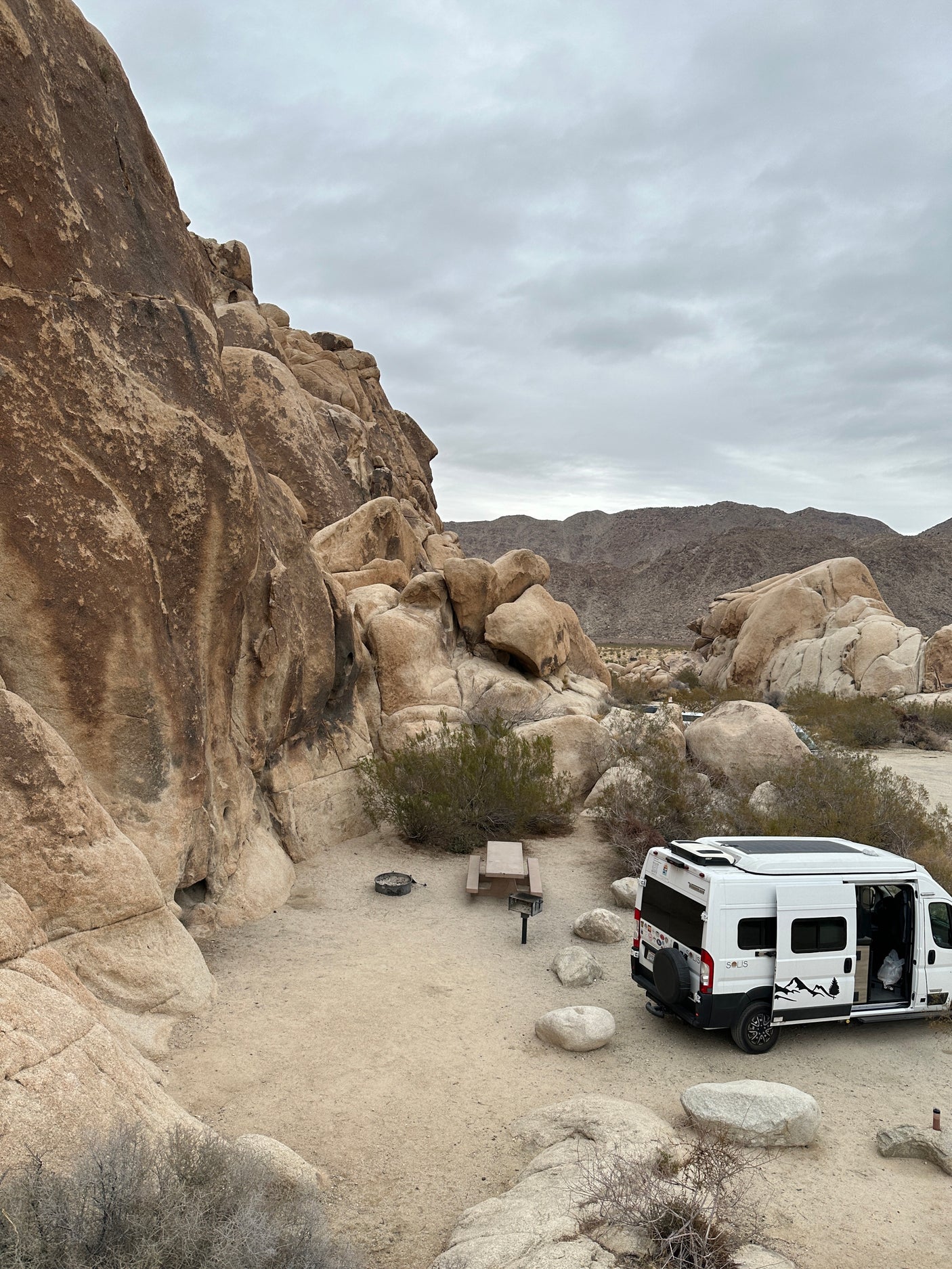 A white RV rental camper van parked at a desert campsite, surrounded by impressive rock formations under a cloudy sky.