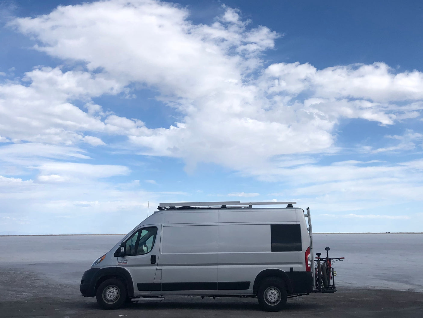 Silver camper van, an RV rental, with bikes on a rack, parked on vast salt flats under a cloudy blue sky.