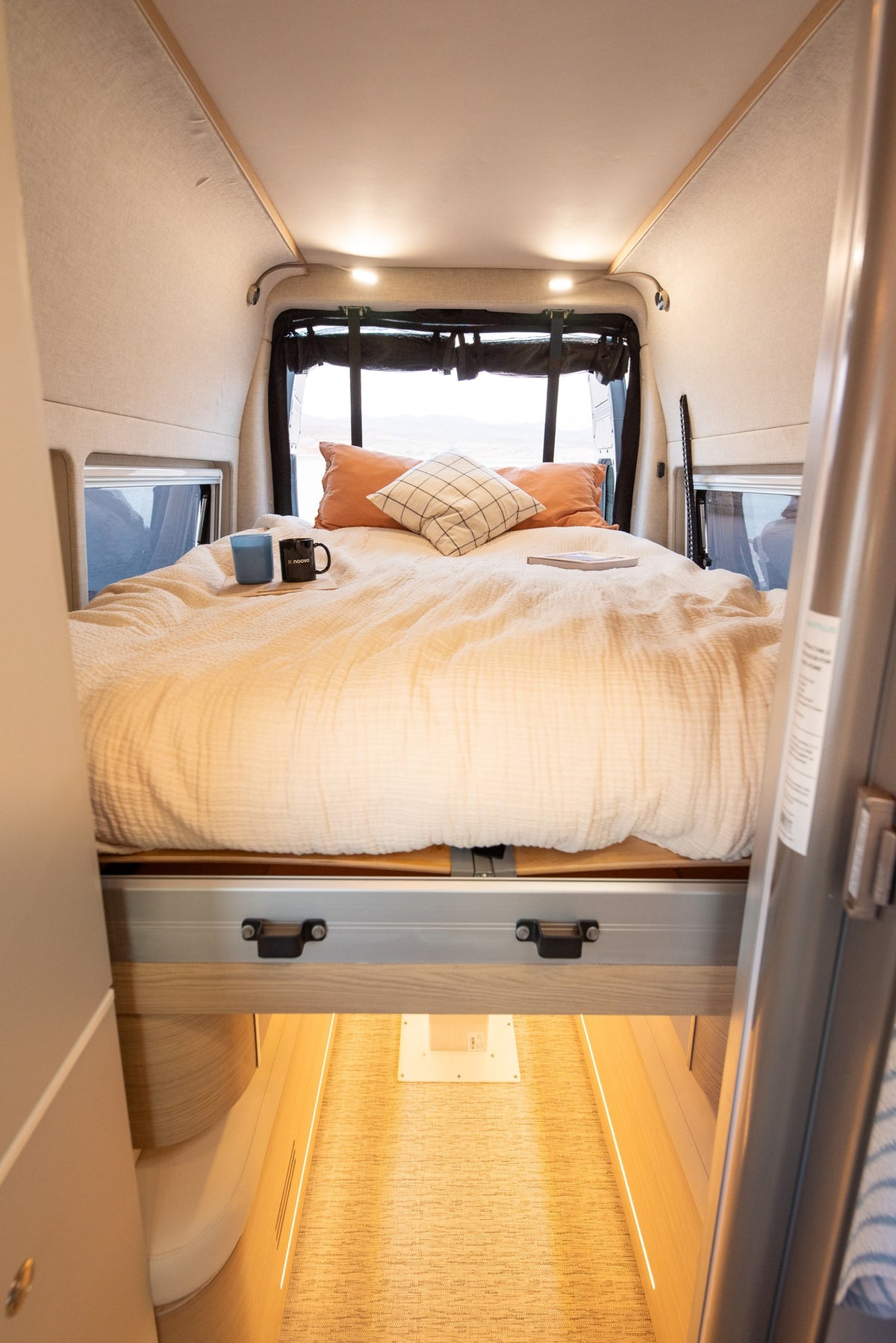 A comfortable, made bed with soft bedding and pillows inside an RV rental, featuring mugs and a book, with windows showing outdoor scenery. — photo 11