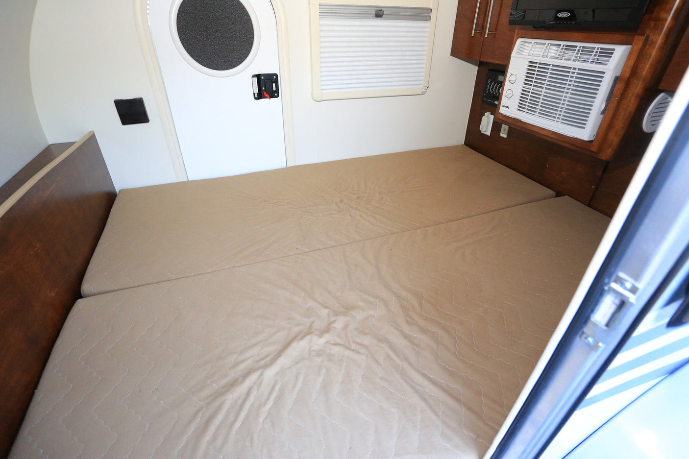 Compact rv rental interior with a light brown bed, white door with circular window, small window with blinds, and a built-in AC unit.