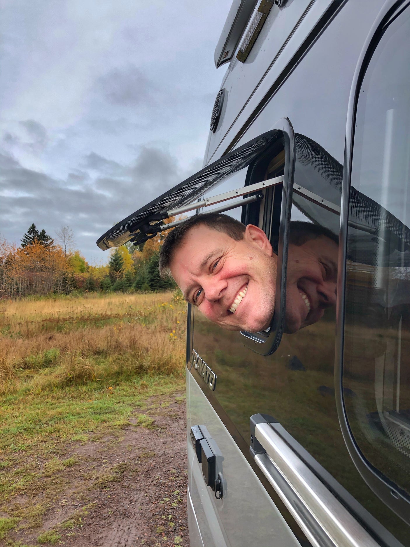 Smiling man and his reflection peek from an RV rental window in a vibrant fall landscape.