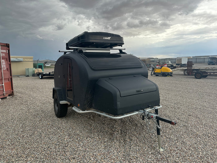 Black off-road teardrop trailer with rooftop box on a gravel lot, available for rv rental.