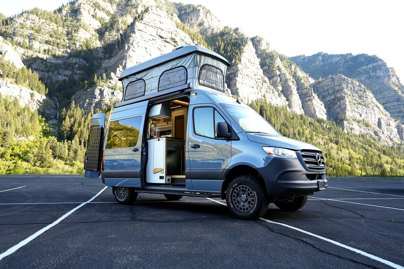 A stunning blue custom Sprinter camper van for rv rental, pop-top raised, parked in a mountain valley lot, ready for adventure.