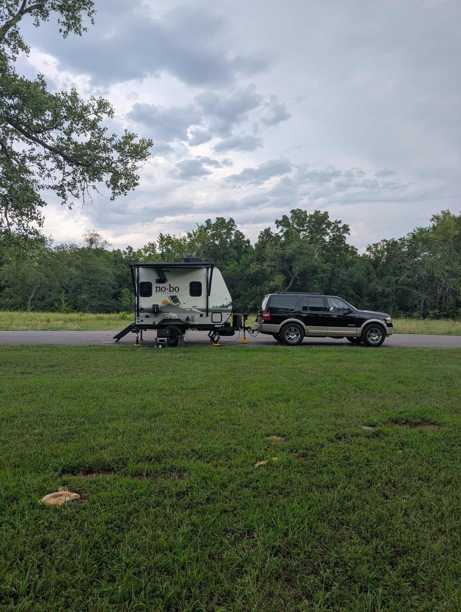 No Boundaries RV rental trailer hitched to a black SUV, set up for camping on a grassy site under cloudy skies.