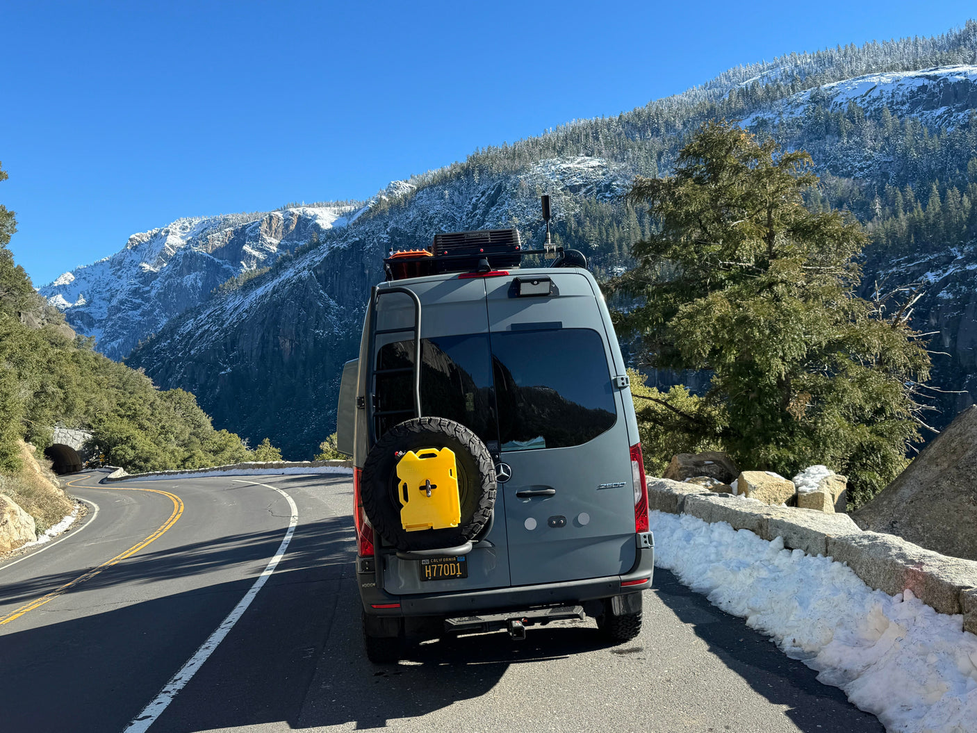 Explore winter wonders in a gray rv rental van on a snowy mountain road, flanked by majestic peaks & a tunnel ahead.