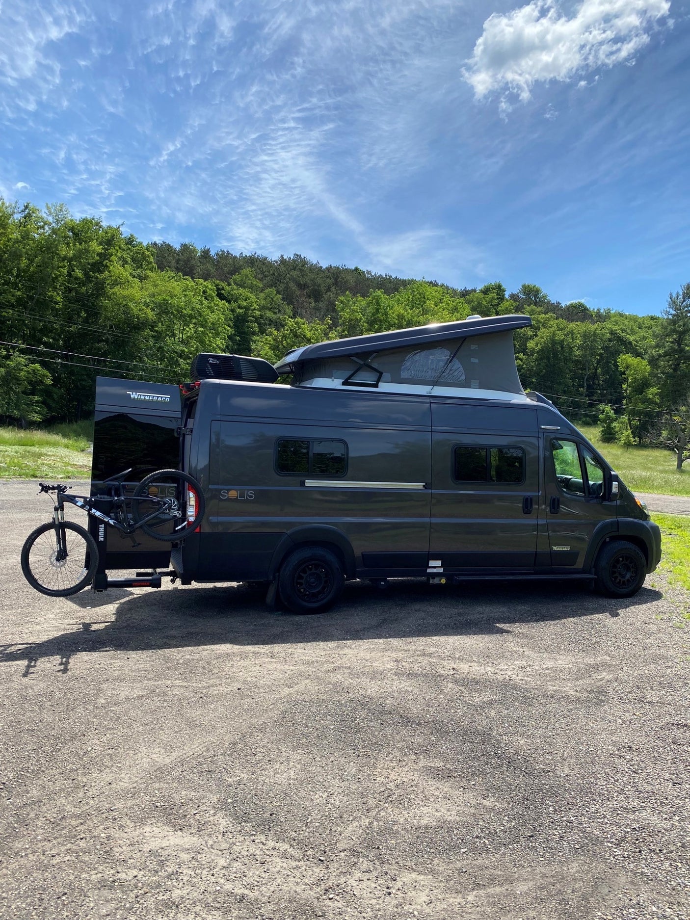 Winnebago Solis RV rental ready for adventure: dark gray van with pop-top, bike rack, parked amidst trees.