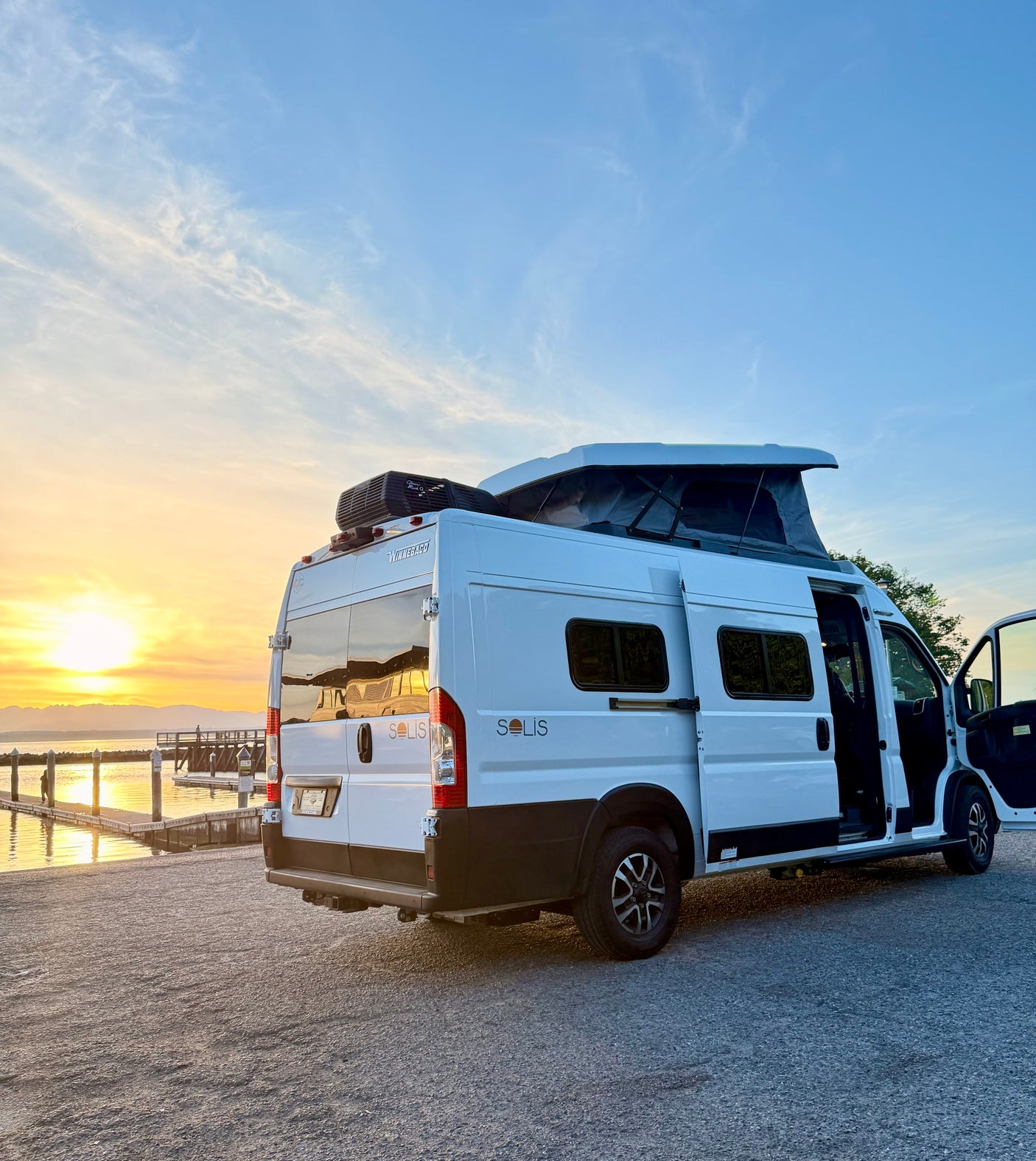 White Winnebago Solis camper van rv rental, doors open & pop-top up, parked by a pier at a scenic sunset waterfront.