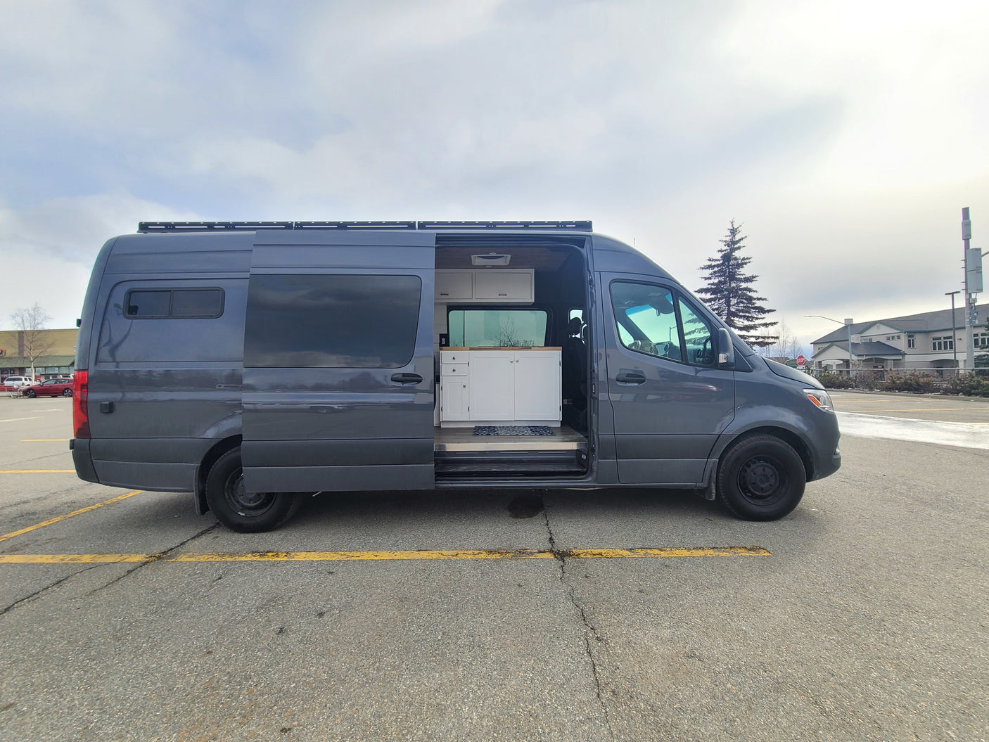A dark grey RV rental van is parked with its side door open, revealing a clean, custom-built interior with white cabinets.