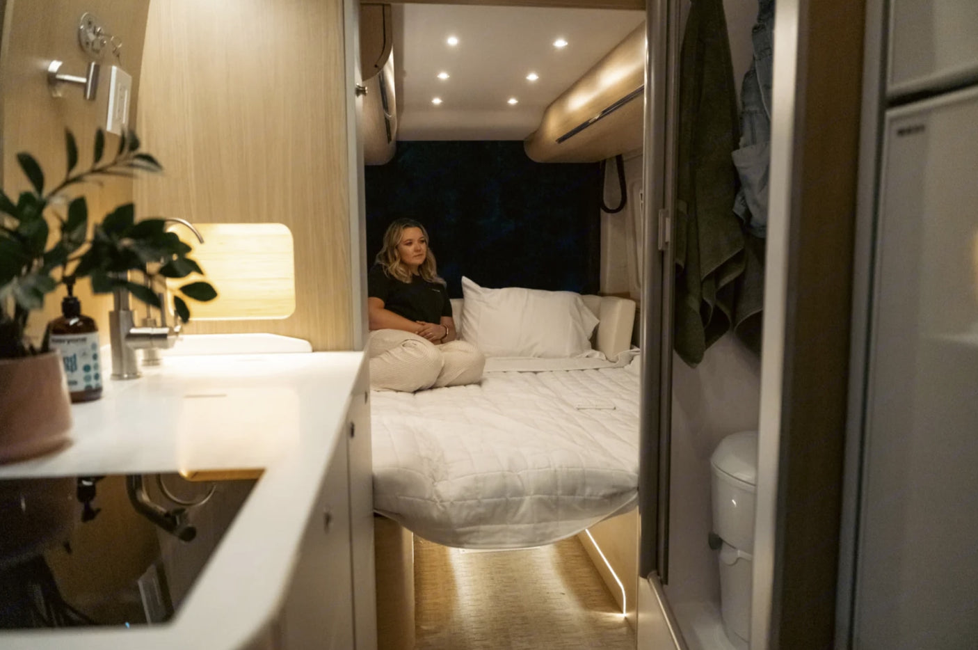 Modern RV rental interior with a woman on the bed, showing kitchen, living, and bathroom areas.