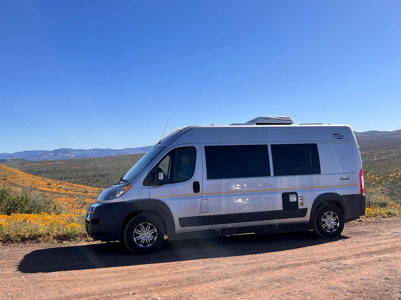 A silver camper van (RV rental) on a dirt road, amidst orange wildflowers with hills under a clear blue sky.