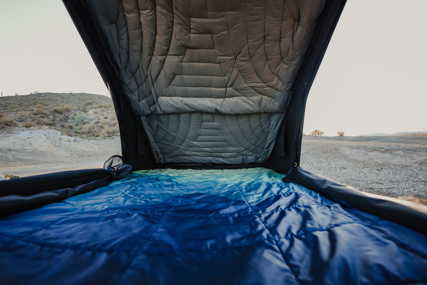 View from inside a cozy RV rental rooftop tent, showing a blue sleeping bag and desert landscape outside. Ideal for camping trips.