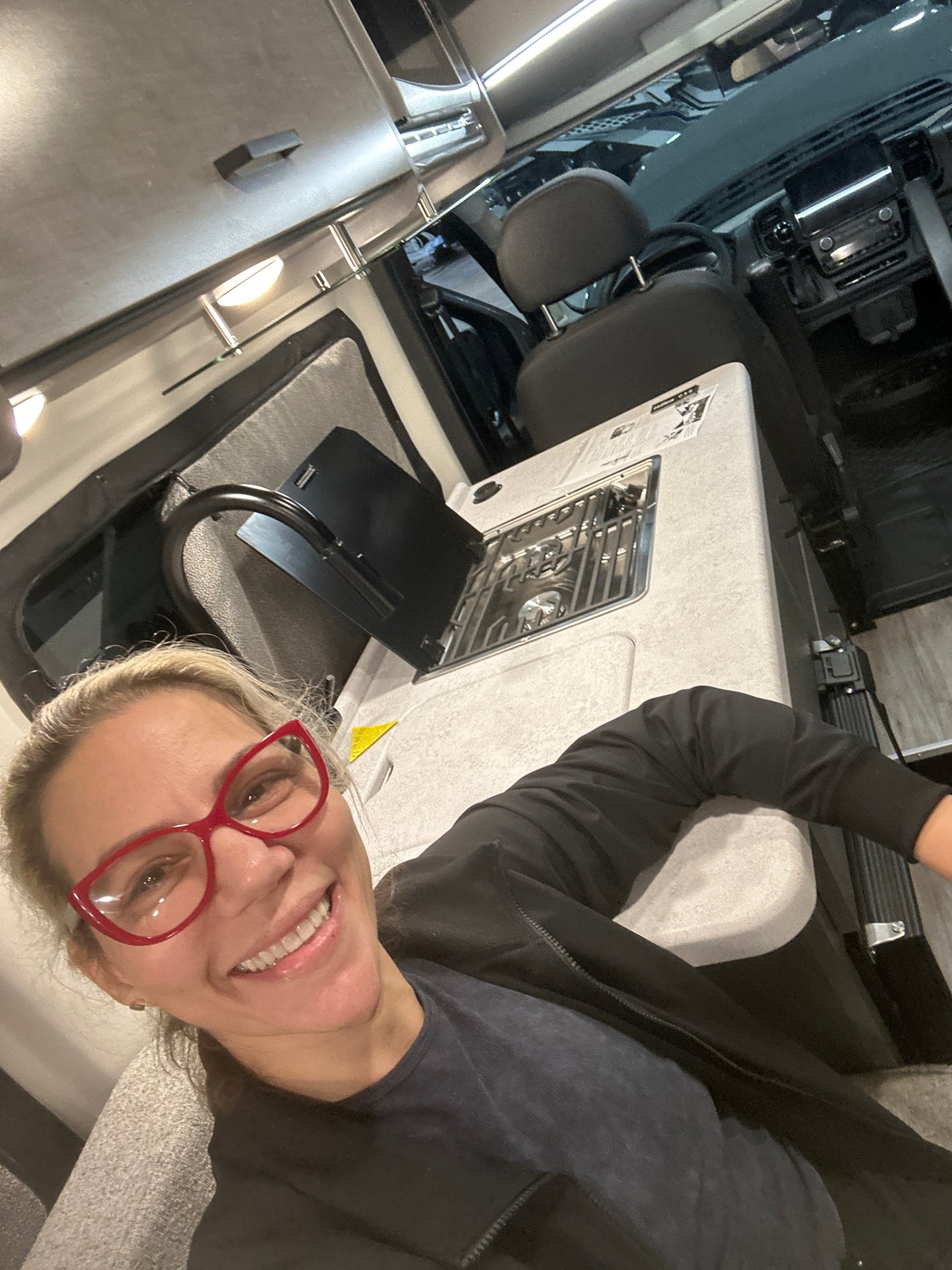 Smiling woman in red glasses inside an RV rental. View from living area shows compact kitchen and front cockpit.