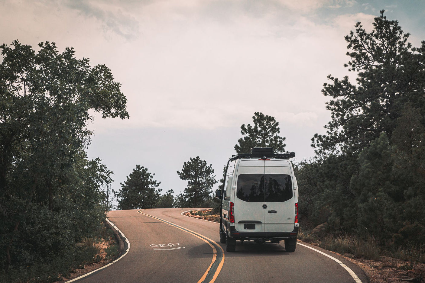 Explore nature with an **RV rental**! A white camper van drives a winding road through a forest under cloudy skies.